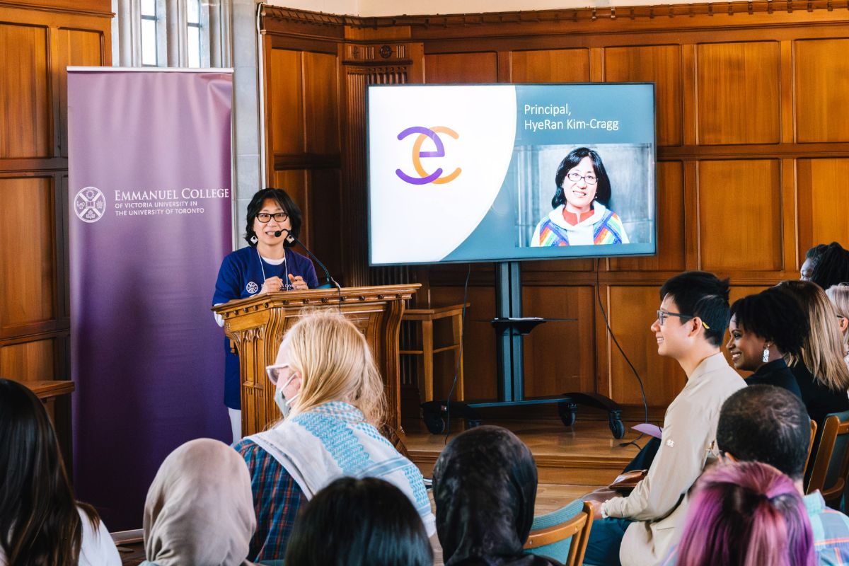 Principal HyeRan Kim-Cragg stands at a podium at the front of the room, speaking to a group of incoming students at Emmanuel College.