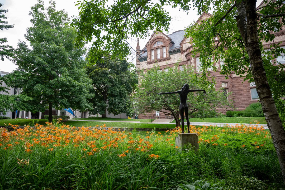 A statue of a crucified woman stands with her arms outstretched, elevated over a bed of vibrant orange flowers and greenery with a red brick building in the background.