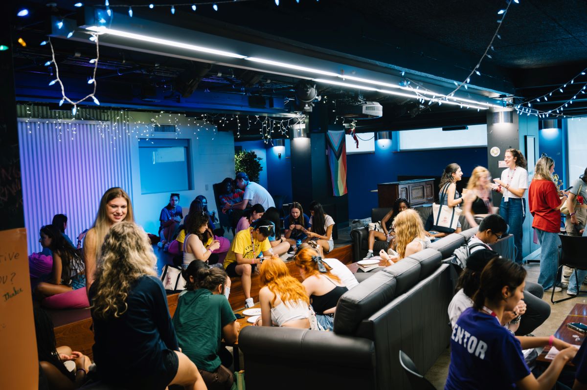 A diverse group of students mingle in a dimmed lounge room, bonding over friendship bracelet making activities.