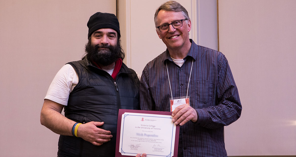 Two men pose for a photo while standing in Alumni Hall holding a diploma. 