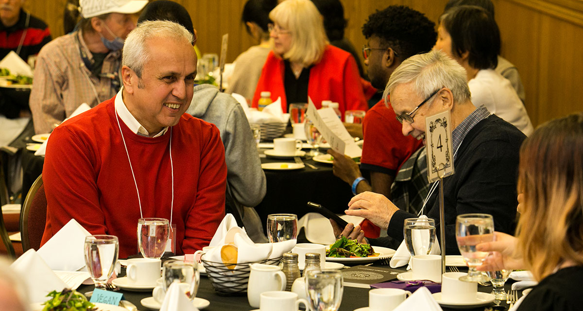  A table of attendees engaged in lively conversations and smiles at Alumni Hall, Victoria University, University of Toronto